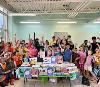 A group of several children and adults raising their fists surrounding a table inside a pool area.