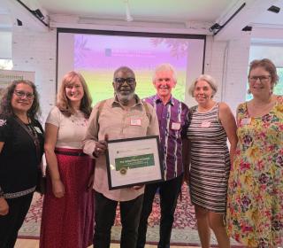 Six people in front of a colourful background, one of which is holding a framed certificate.