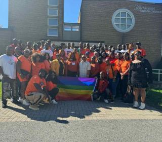 A group of African people, many wearing orange shirts, holding up a pride flag outside of Kitchissippi United Church.