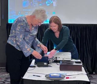 Two women set up electronic equipment on a boardroom table.