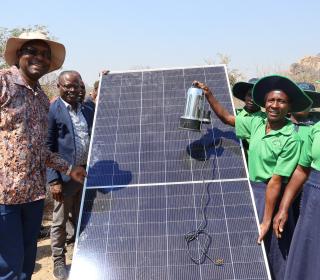 A group of people pose surrounding a solar panel outdoors in Zimbabwe.