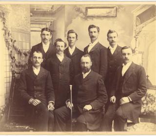 A group of men in 1892, in the formal dress of the time, pose as a group, with the front central figure holding an engraved wooden staff.