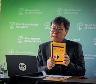 A man in mid-speech holds up a book, sitting down before a laptop with a United Church sticker on it at a desk.