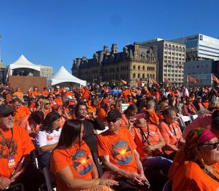 A large crowd of people wearing orange shirts sitting outside of the Canadian Parliament buildings.