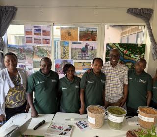 A group of individuals smiling in front of a table holding produce and grains, with photos of farming in action behind them