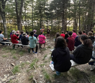 A group of kids sit on benches outdoors in a forest setting