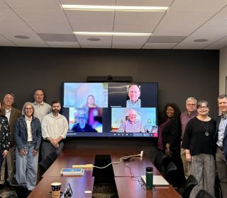 Nine people pose in a boardroom around a tv screen featuring more smiling in an online meeting