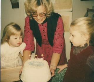 Two young girls celebrate a birthday with cake held by an adult woman in the 1980s