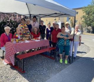A group of a dozen stand around a brightly decorated picnic table outdoors