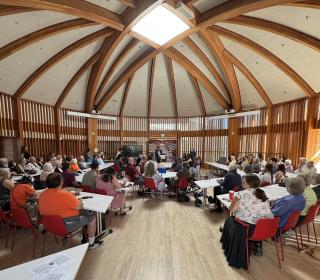 Several dozen people gather at desks in a large room with vaulted timber ceilings.