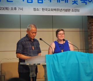 A Korean person and white person speak at a podium with a banner in Korean behind them