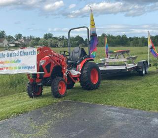 A rainbow picnic table is carried on a trailer by a tractor bearing a "Sackville United Church" banner