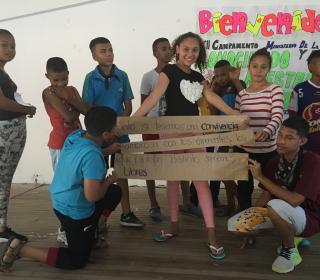 Colombian children and teenagers participate in a theatre education program. The stand in a group in front of a sign that has "Bienvenidos" (welcome).