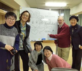 Kyofukai staff (four Japanese women) and United Church representatives (a woman and man) gather about an easel for a photo.