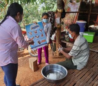 Two women wearing masks hold a sign showing proper hand hygiene during COVID-19 while a seated man wearing a mask demonstrates handwashing.
