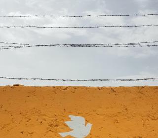 A wall painted with a dove topped by barbed wire against a cloudy sky.