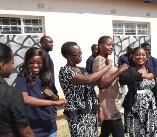 A group of six young women in Zambia each flex their right arm, while a group of young men stand behind them.