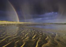 A rainbow seen from a beach while storm clouds swirl overhead.