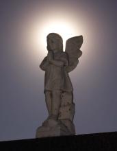 A white marble statue of an cherub-like angel silhouetted by a halo of the sun.