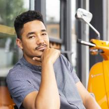 A casual portrait of Kofi Hope, a young Black man, sitting outside near a motor bike.