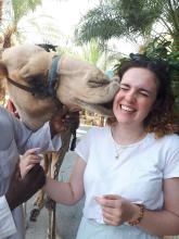 A young woman gets a big sloppy kiss from a camel on the side of her face and squirms.