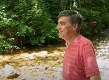 Alf Dumont, an Indigenous man, stands beside a creek, smiling and looking upstream.