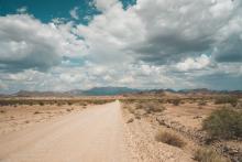A dirt road running straight through the sparse desert, with dramatic clouds overhead.