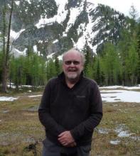 Rev. David Boyd, a man with a white beard, wearing dark glasses, in a British Columbia valley surrounded by snow dappled mountains.