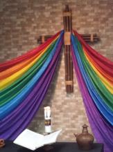 A cross made of bamboo branches hangs on a chapel wall draped in rainbow-coloured cloth.