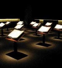 A dark room, except for spotlights shining on a number of individual lecterns, each containing an open book which holds the testimonies of the victims of Okinawa.