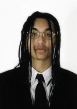A young Black man looking classy in a suit and tie with locks and golden glasses.