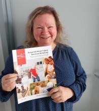 The author, Rev. Shelley Pick, a woman with light brown hair, twinkling eyes, smiles while holding a book from Brain Injury Nova Scotia.