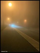 A moody photograph of a foggy street at night, with street lights dimly showing through and the headlights of a car approaching in the darkness.