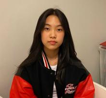 A Korean-Canadian teenage woman sitting and looking directly at the camera, wearing a red and black windbreaker.