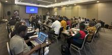 A packed conference room lined with people writing or on laptops. Four presenters sit at the front.