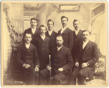 A group of men in 1892, in the formal dress of the time, pose as a group, with the front central figure holding an engraved wooden staff.