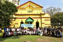 A large group photo of Indian students and educators outside of a yellow building