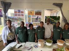 A group of individuals smiling in front of a table holding produce and grains, with photos of farming in action behind them
