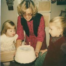 Two young girls celebrate a birthday with cake held by an adult woman in the 1980s