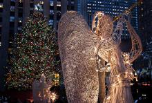 A sculpture of an angel blowing a horn, standing in front of a Christmas tree with multi-coloured lights.