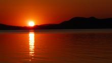 A picture of a sunset over water with mountains in the background. A deep red-orange colour saturates the evening scene.
