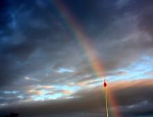 A photo of a Canadian flag on a flagpole apparently being found at the foot of a rainbow, against the contrast of a dramatic dark sky.