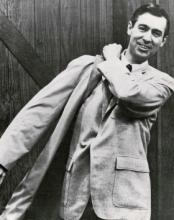 A black and white photograph of Fred Rogers taken in the late 1960s. He is a White man with dark hair and an inviting smile. He is shown wearing a light coloured suit jacket while pulling a bag over his shoulder.