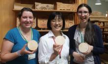 Three young women who were participants in the Minority Youth Forum hold a wooden carving they did. The White Canadian authors of the blog post, Martha and Sarah are on the right and left, with Miyauchi from Japan is between them. They are all smiling.