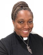 A portrait of Rev. Dr. Karen Georgia Thompson, a Black woman with beautiful braids atop her head in a bun, wearing a clergy collar and a metal owl pendant.
