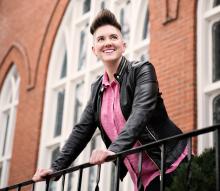 A photo of writer, speaker, musician, and atypical activist Pam Rocker, a person with a black leather jacket, pink shirt, and pompadour hair-do smiling brightly on the steps of church.