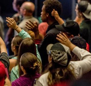 A crowd of young people in an auditorium lifts their right arm in the air