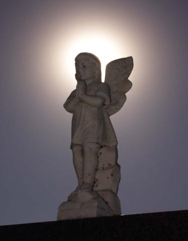 A white marble statue of an cherub-like angel silhouetted by a halo of the sun.