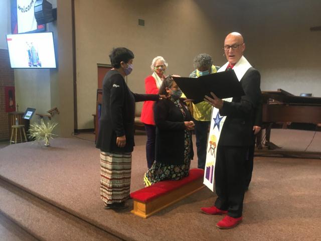 Deb Anderson-Pratt kneels at ordination ceremony while minister reads and woman presents her with eagle feather