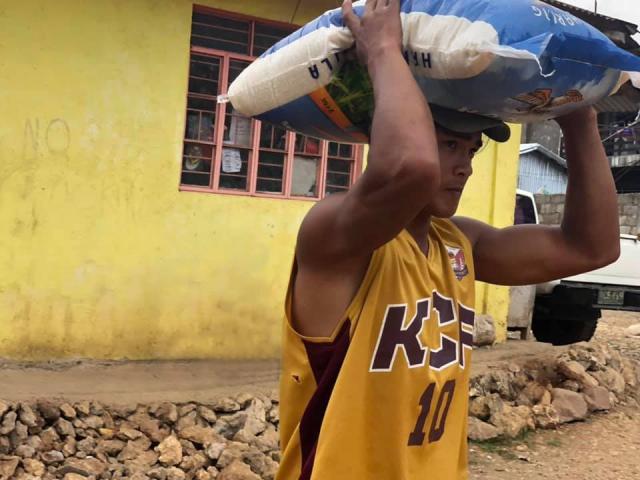 A man carries a large bag of rice and a yellow house is in the background.